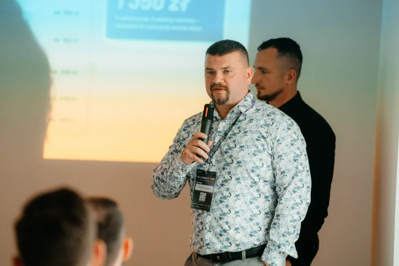 Michał Olejarski z mikrofonem odpowiada na pytania uczestników, za nim Radosław Zacha