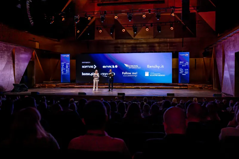 Prowadzący na scenie na tle ekranu z logotypami partnerów konferencji, wśród nich General Informatics