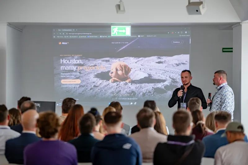 Strona Aizor wyświetlona na ekranie podczas warsztatu: piesek wyglądający z krateru na Księżycu i nagłówek „Houston, mamy przyjaciela.”; obok Radosław Zacha z mikrofonem i Michał Olejarski