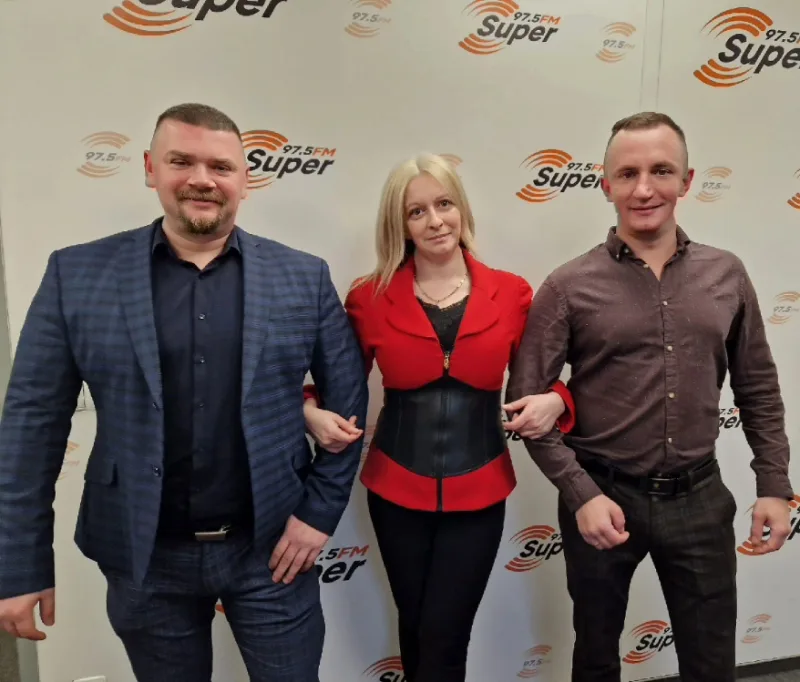 Trzy osoby stoją pod rękę na tle ścianki z logo Radia Super FM 97,5 FM: od lewej Michał Olejarski w granatowej marynarce, Monika Różycka w czerwonym żakiecie i Radosław Zacha w brązowej koszuli